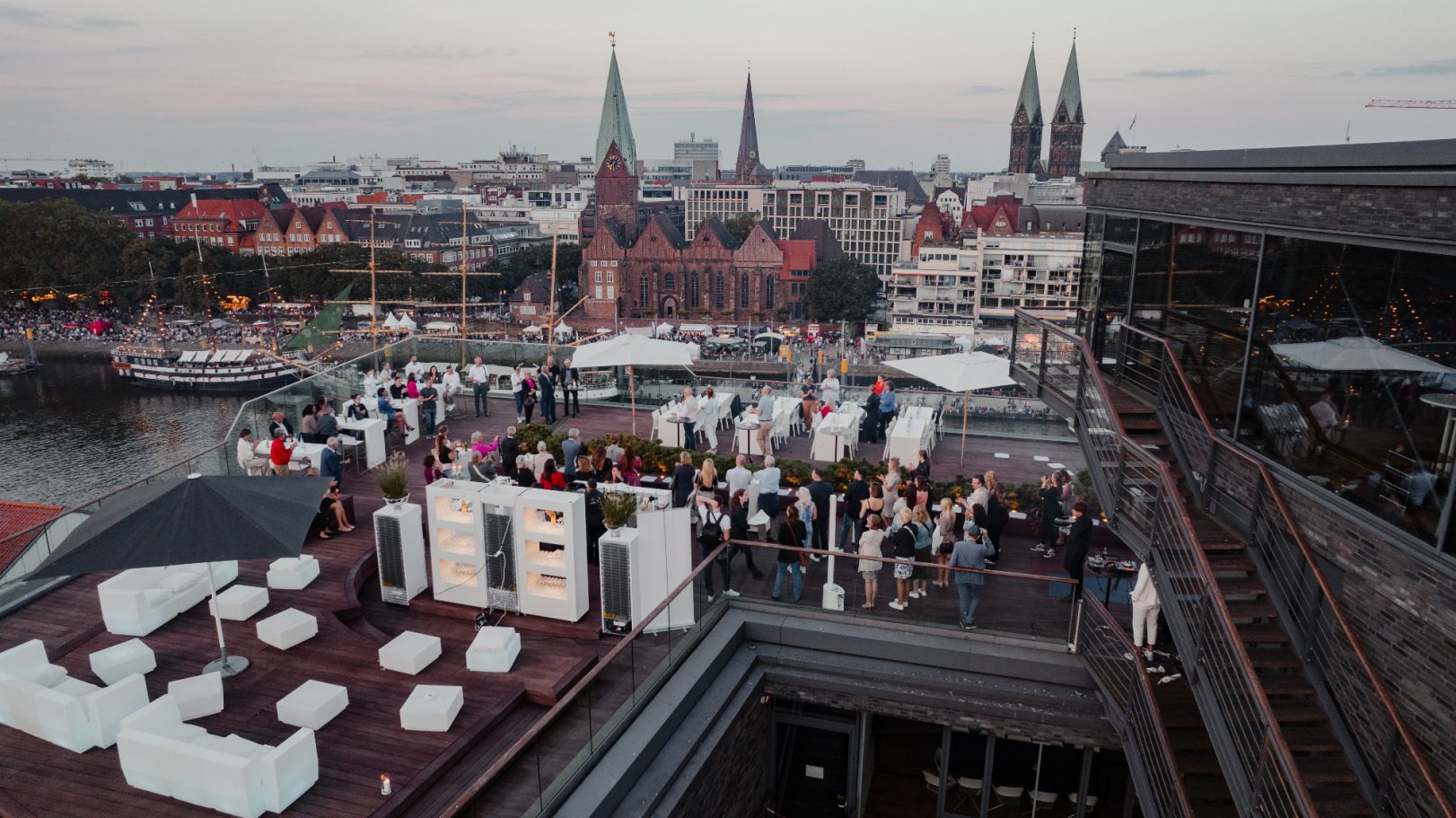 Blick von oben auf eine lebhafte Dachterrasse mit Stadtansicht und feiernden Menschen.