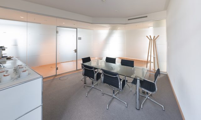 Moderner Konferenzraum mit Glastisch, schwarzen Stühlen und heller, minimalistisch eingerichteter Wand.