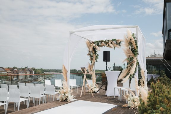 Zelt mit Blumenarrangements für eine Hochzeit, Stühle davor, Blick auf Wasser im Hintergrund.