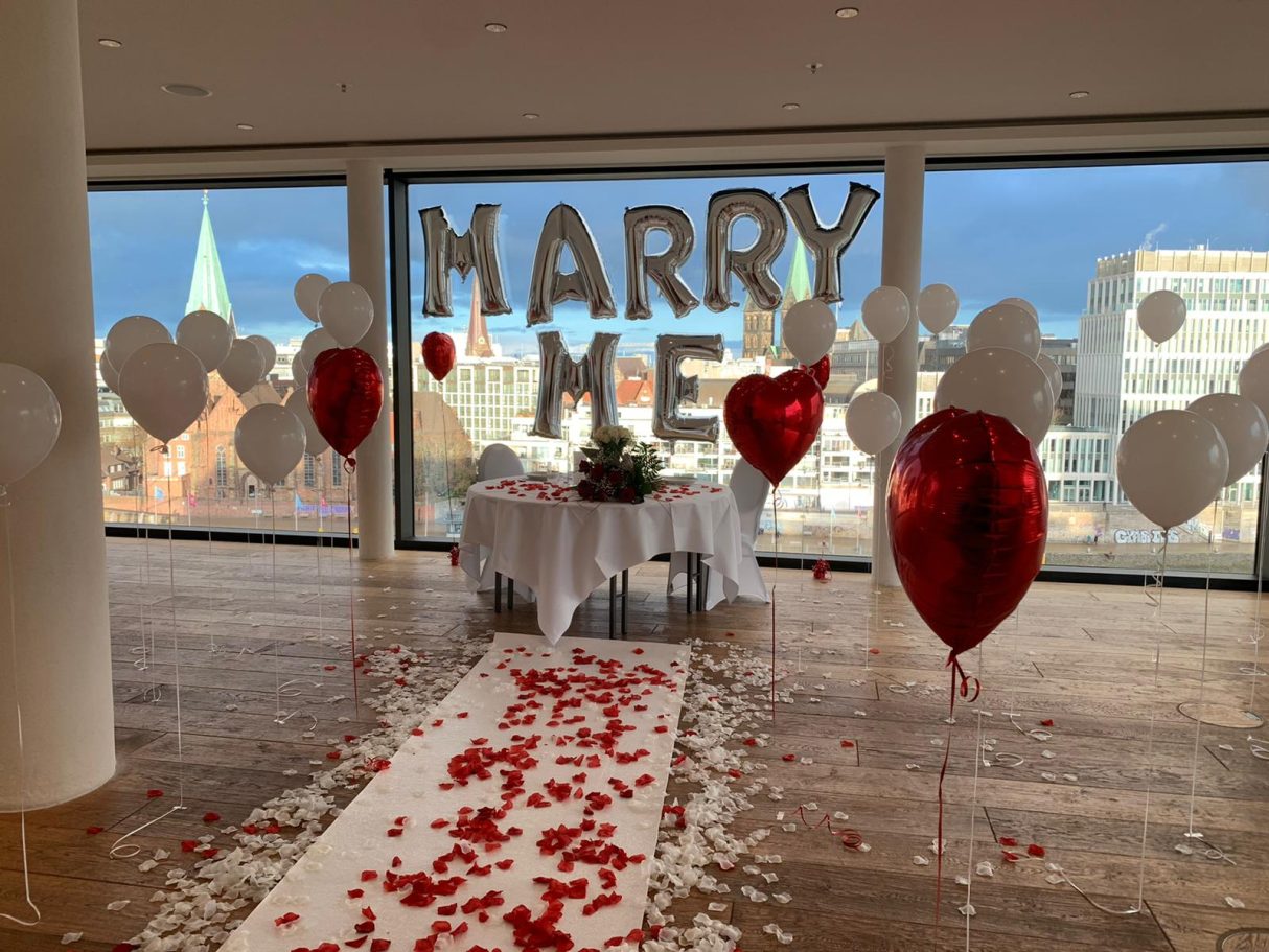 Tisch mit "MARRY ME"-Dekoration, Herzballons und Rosenblättern für einen Heiratsantrag.