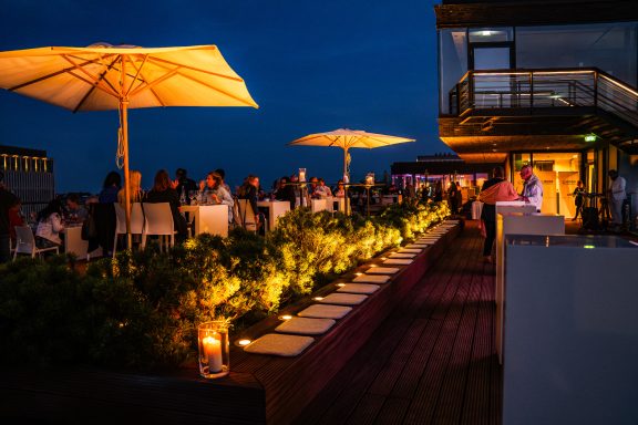 Beleuchtete Dachterrasse mit Essbereich und Sonnenschirmen bei Abenddämmerung.