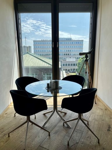 Ein runder Glastisch mit vier schwarzen Stühlen, Blick auf ein Gebäude und ein Fernrohr.